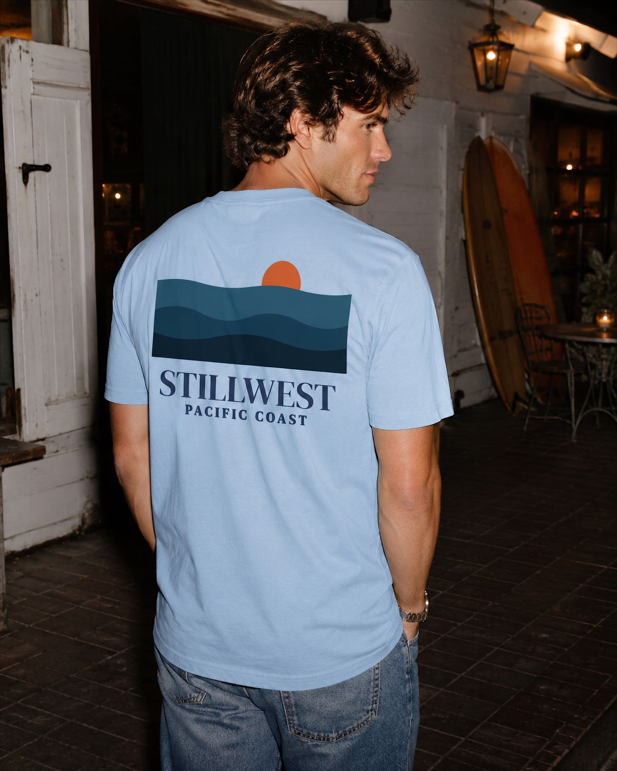 A man wearing a light blue t-shirt with a graphic of layered blue waves, an orange sun, and the text 'STILLWEST PACIFIC COAST' on the back, standing in a dimly lit outdoor patio setting.