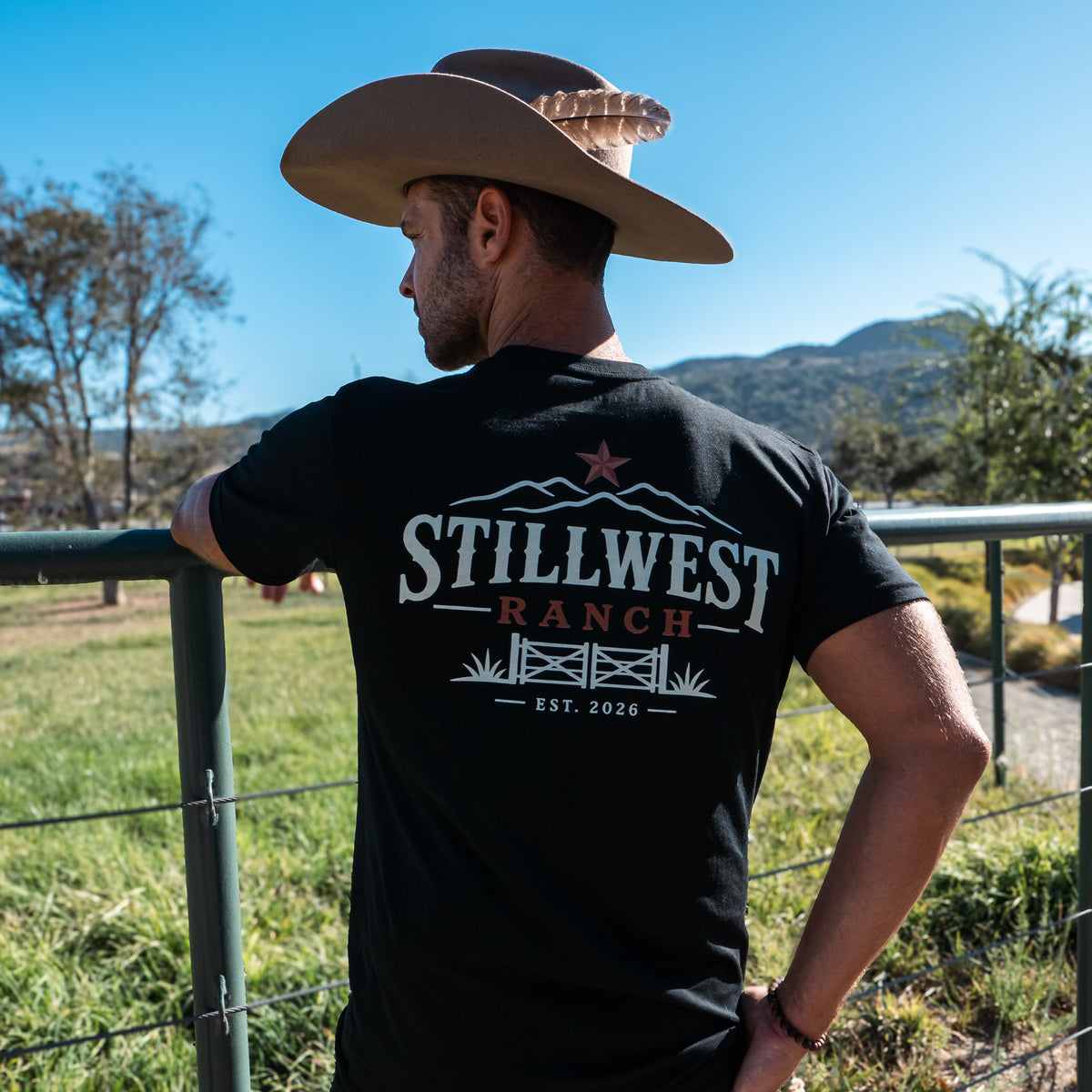 A man wearing a brown cowboy hat and a black t-shirt with a 'Still West Ranch' graphic on the back, leaning on a metal fence in an outdoor ranch setting with mountains and blue sky in the background.